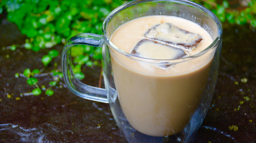 clear mug of iced coffee on pavement with greenery