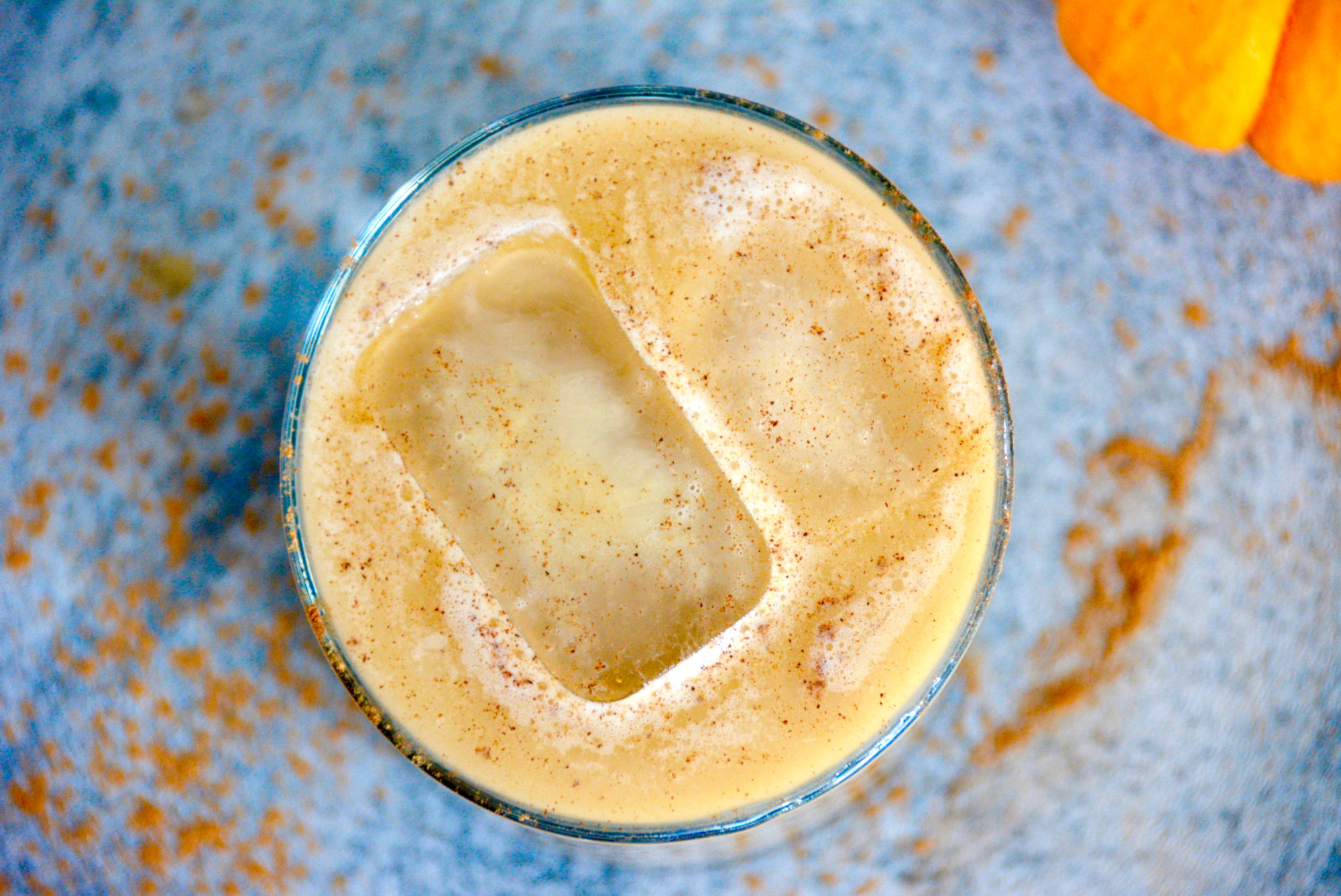 from top - view of pumpkin spice latte in a high ball glass with a blue background a small pumpkin and two ice cubes in the latte