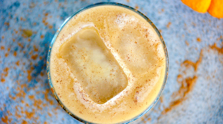 from top - view of pumpkin spice latte in a high ball glass with a blue background a small pumpkin and two ice cubes in the latte