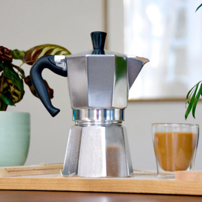 bialetti moka pot next to plants and a cup of coffee on wood table