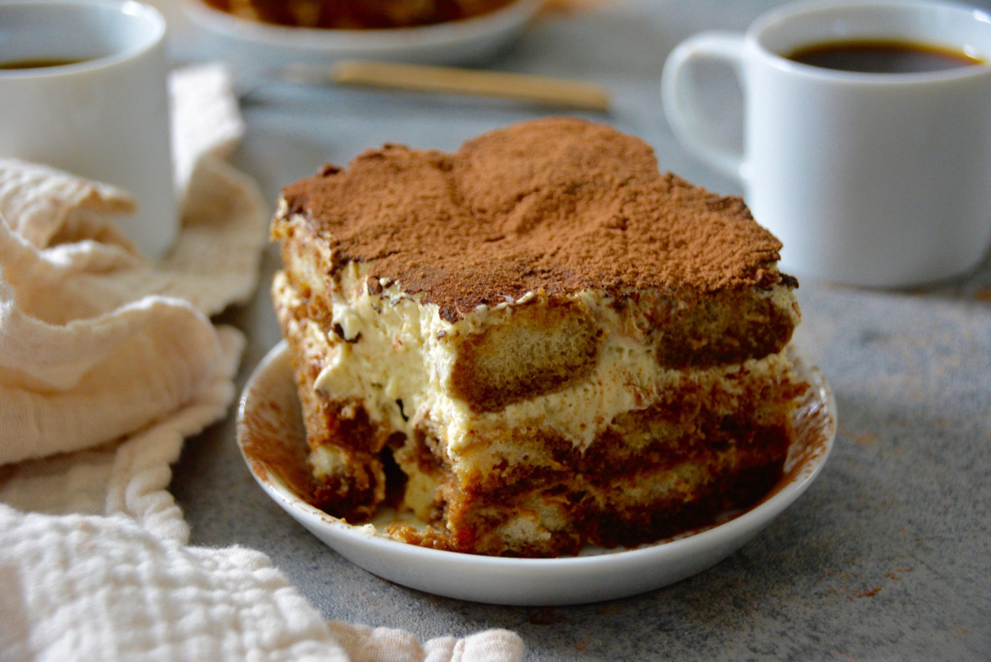 tiramisu on a plate with coffee cups in background